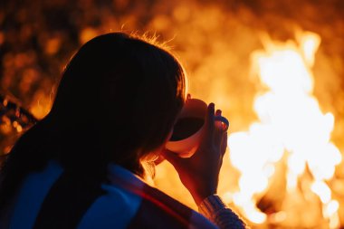 Şenlik ateşinin yanında oturmuş sıcak çay ya da kahve içen bir kızın fotoğrafı. Doğada vakit geçirmek, şenlik ateşi yakmak.