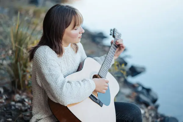 Young beautiful girl playing the guitar near the river. Girl practicing playing the guitar, looking on the water