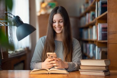 Mutlu gülümseyen üniversite öğrencisi kız cep telefonunu kullanıyor. Üniversite kütüphanesinde çalışan, kitapları ve telefonuyla masada oturan bir öğrenci.