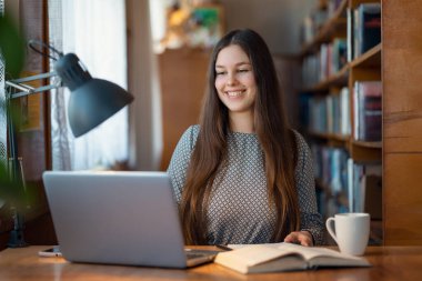 İnternetten online ders alan gülümseyen kız. Üniversite kütüphanesindeki neşeli kız öğrenci, ödevini hazırlıyor.