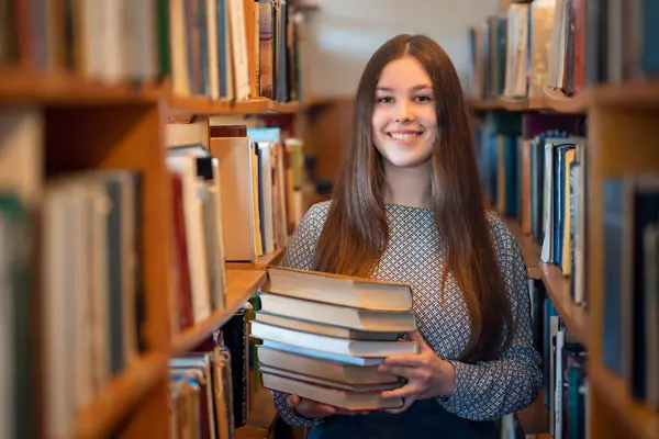 Kütüphanede kitapların arasında gülümseyen genç kız. Mutlu kız üniversite ödevlerine hazırlanıyor, edebiyata bakıyor.