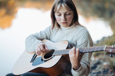 Nehir kenarında gitarla müzik çalıyordu. Genç kadın sonbaharda serin bir akşamda enstrümanla gölün kıyısında oturuyor.