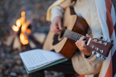 Kamp ateşinin yanında doğada pratik yapan bir kız gitariste yakın çekim. Bir kızın notla gitar çalıp şenlik ateşinin yanında yeni bir şarkı bestelediği sıcak bir akşam fotoğrafı.