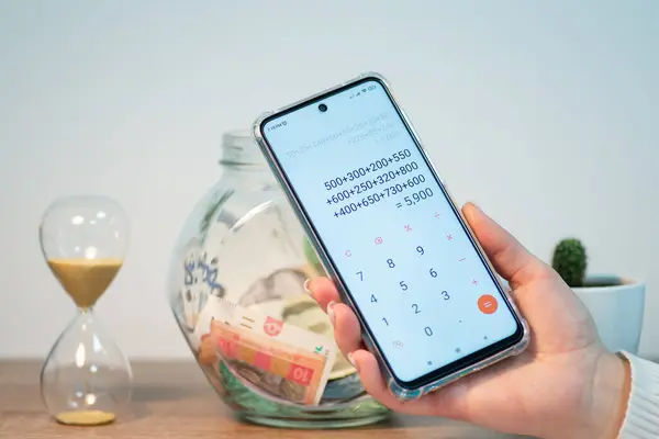 Counting saved money for a purchase. Phone with calculator in female hands with jar with saved up money on the background