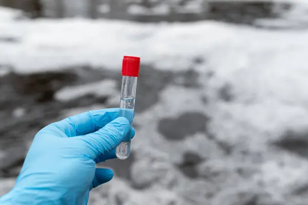 Test tube with red cap and water inside in male hand in blue medical gloves, close up. Foaming dirty water on the background, concept of water pollution