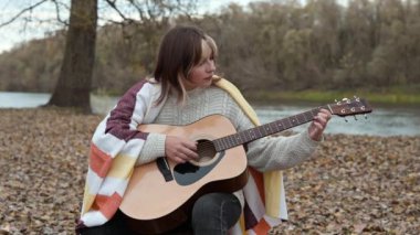 Nehir kıyısında gitar çalan genç bir kız. Ekoseyle kaplı güzel bir kız sonbahar akşamında doğada akustik gitar çalıyor.