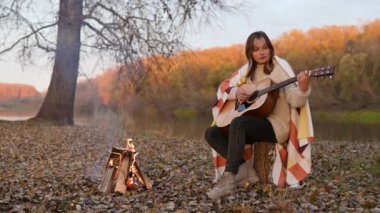 Yanlız kız ateşin başında oturmuş gitar çalıyor. Ormanda kamp, sonbahar parkı, doğada boş zaman, yalnızlık.