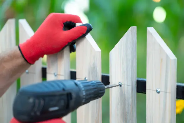 Eldivenlerini çıkarıp elektrikli tornavida yardımıyla ahşap çit yapıyor. Ev entrikalarını kendin düzenlemek, kendi ekipmanlarınla yeni çitler yapmak.
