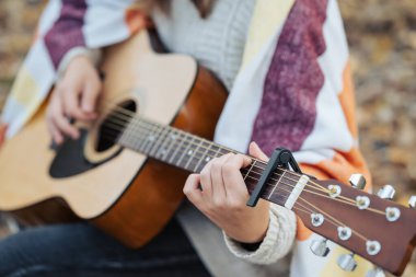 Sonbahar doğasında gitar çalan genç bir kadının yakın çekimi. Elinde gitarla, sıcak ekoseyle gezen, sonbahar ormanlarında gitar çalan, Capo kullanan bir kız.