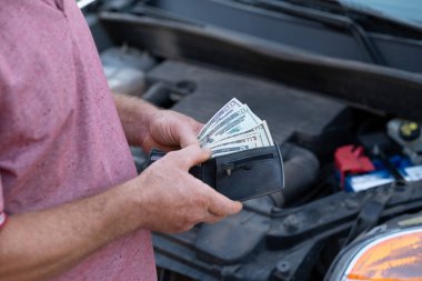 Adam araba tamiratlarını kararlılıkla değerlendirirken dolarları elinde tutuyor. Bir adam arabasının motorunu bir tomar parayla kontrol eder, evinin önünde tamir masraflarını ve çözümleri düşünür.