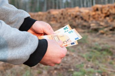 Ormansız bölgede avroluk banknotlar saymak. Avro notlarını istiflenmiş kütüklerin arka planına karşı tutan eller, Avrupa 'nın orman tahribatına katılımını simgeliyor