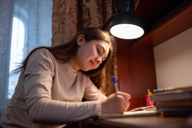 Evde çalışırken gülümseyen genç bir kız. Neşeli ve motivasyonlu bir masada ödev yapan mutlu genç kız.