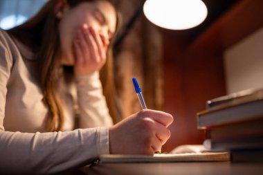 Çalışırken elinde kalem tutarken esneyen uykulu genç kız. Genç kız ödevlerini yaparken esniyor. Yorgunluk, rutin ve geç çalışma kavramı