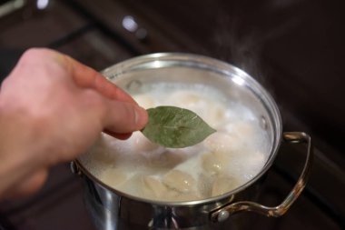 Kaynayan pelmeni 'ye el yaprağı ekliyorum. Yakın plan bir el, taze bir defne yaprağını kaynamış bir tencereye koyuyor ve hamur köftesi pişiriyor. Yemek yaparken lezzeti arttırıyor.