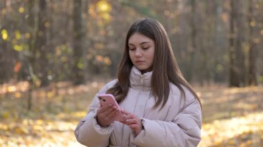 Genç kız sonbahar ormanlarında üzücü mesajlar okur. Genç kız, sonbahar ormanlarında yürürken hüzün ve duygusal baskı gösteren telefon mesajlarını okur.