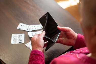 Boş cüzdana bakan yaşlı bir kadın. Masasında kullanılmış ilaç paketleri olan boş cüzdanı olan yaşlı bir kadın, yoksulluğu ve karşılanamaz sağlık hizmetlerini simgeliyor.