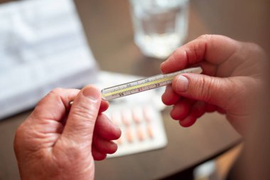 İlaçlar arasında yaşlı bir kadının elindeki termometre. Üst düzey kadın ellerini termometreyle yakın plan, arka planda haplar olan masa, evde sağlık kontrolü gösteriyor.