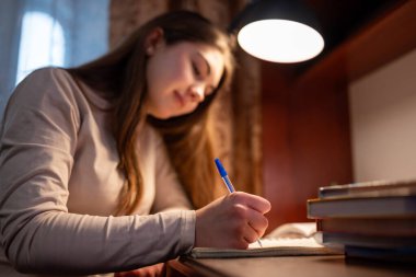 Akşamları ödev yapan liseli kız. Genç bir kız elinde lambayla masada oturuyor, kalemle deftere yazıyor, ödevlerine konsantre oluyor.