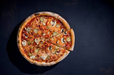 Appetizing pizza with hunting sausages, mozzarella cheese, pickled cucumbers, chili peppers on a tomato base on black background.