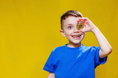 Mavi tişörtlü küçük mutlu anaokulu çocuğu sarı arka planda gözlerinin önünde Bitcoin tutuyor..