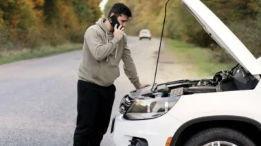 Genç bir adam beyaz kırık bir arabanın yanında kaputu açık duruyor ve yardım istiyor..
