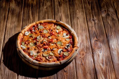 Appetizing pizza with hunting sausages, mozzarella cheese, pickled cucumbers, chili peppers on a tomato base on wood background.