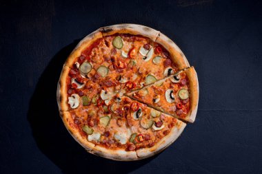 Appetizing pizza with hunting sausages, mozzarella cheese, pickled cucumbers, chili peppers on a tomato base on black background.