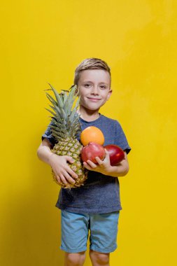 Sarı arka planda taze ananas, elma ve portakal tutan gülümseyen çocuğun stüdyo fotoğrafı. Sağlıklı bebek maması konsepti.