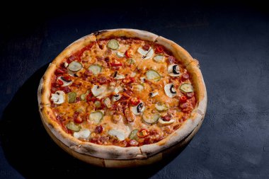 Appetizing pizza with hunting sausages, mozzarella cheese, pickled cucumbers, chili peppers on a tomato base on black background.