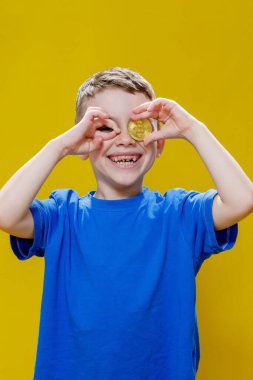 Mavi tişörtlü küçük mutlu anaokulu çocuğu sarı arka planda gözlerinin önünde Bitcoin tutuyor..