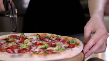 Freshly baked appetizing pizza with cherry tomatoes, mushrooms, broccoli, red onion on a tomato base cuting into slices with a pizza cutter for serving in a pizzeria.