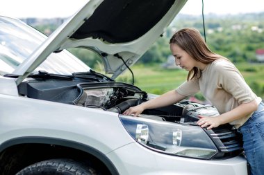 Arabanın kaputu açık duran kafası karışmış kadın sürücü bozuk motora bakıyor.