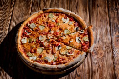 Appetizing pizza with hunting sausages, mozzarella cheese, pickled cucumbers, chili peppers on a tomato base on wood background.