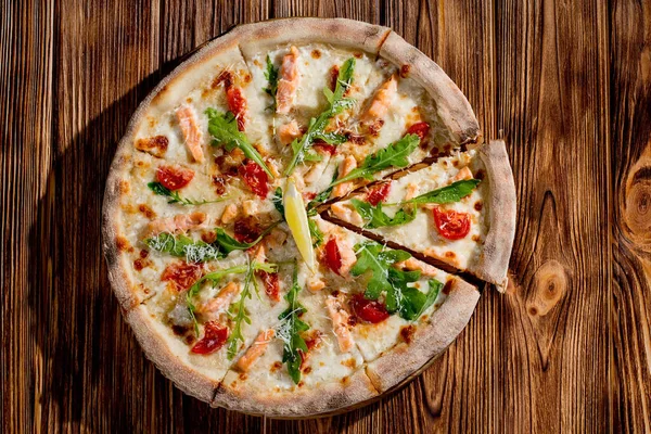 Pizza with salmon, mozzarella, cherry tomatoes, arugula, lemon and parmesan. Italian cuisine on a woden background.