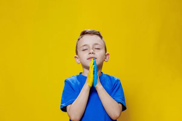 Ukrainian boy prays for Ukraine. Children against war. A boy in a blue T-shirt on a yellow background folded his hands painted in the Ukrainian flag for prayer.