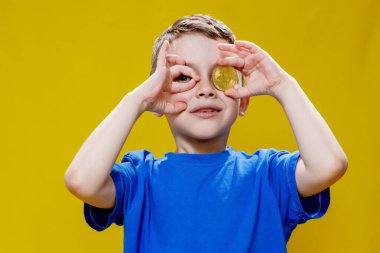 Mavi tişörtlü küçük mutlu anaokulu çocuğu sarı arka planda gözlerinin önünde Bitcoin tutuyor..
