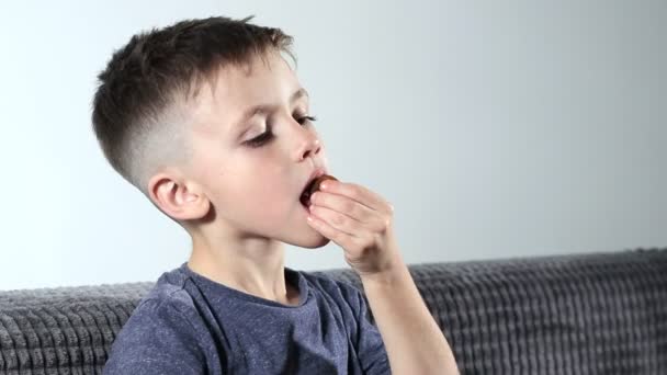 一个小男孩坐在舒适的沙发上 打开糖果 嘴里衔着番石榴 小孩子喜欢吃糖果 — 图库视频影像