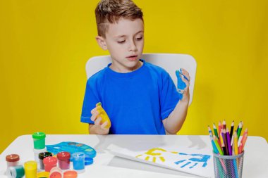 Küçük bir anaokulu öğrencisi masada oturuyor, avuç içlerini bir kağıt parçasına çiziyor. Renkli el izleri. Anaokulu geliştirme.