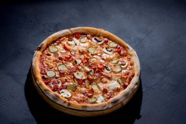 Appetizing pizza with hunting sausages, mozzarella cheese, pickled cucumbers, chili peppers on a tomato base on black background.