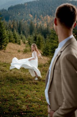 The bride is circling in her wedding dress, and the groom is looking at her. Wedding photo session in the mountain.