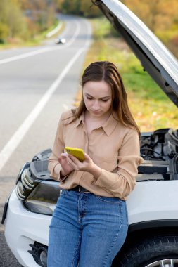 Bir kadın yol kenarında arabasının yanında yardım bekliyor. Kaputu açık, kırık arabalı bir kadın. Bozuk arabanın yanında cep telefonu olan üzgün genç bir kadın..