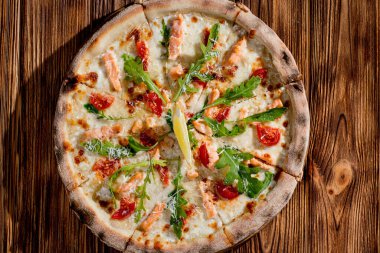 Pizza with salmon, mozzarella, cherry tomatoes, arugula, lemon and parmesan. Italian cuisine on a woden background.
