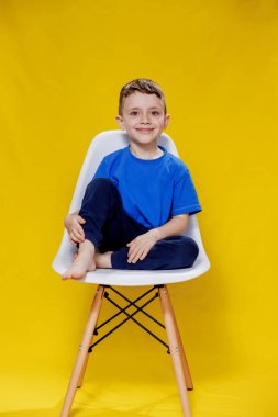 Cheerful little fair-haired boy in blue t-shirt and pants sitting on a chair and posing on yellow background.