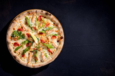 Pizza with salmon, mozzarella, cherry tomatoes, arugula, lemon and parmesan. Italian cuisine. On a black background. Free space for text. View from above