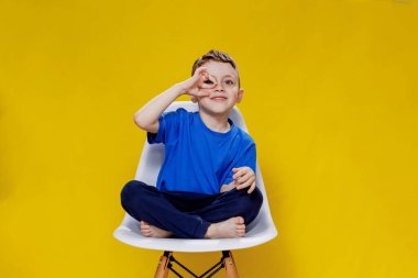 Cheerful little fair-haired boy in blue t-shirt and pants sitting on a chair and posing on yellow background.