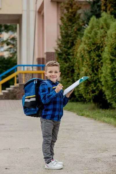 学校の庭でノートにメモを書いて笑顔で バックパックとカジュアルな服を着た幸せな少年 — ストック写真