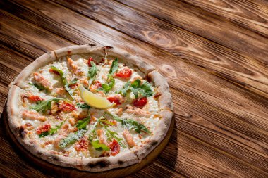 Pizza with salmon, mozzarella, cherry tomatoes, arugula, lemon and parmesan. Italian cuisine on a woden background.