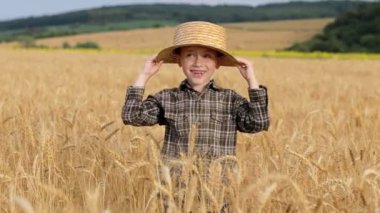 A smiling little farmer boy in a plaid shirt and straw hat poses for a photo in a wheat field. Heir of farmers.