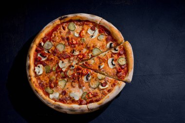 Appetizing pizza with hunting sausages, mozzarella cheese, pickled cucumbers, chili peppers on a tomato base on black background.