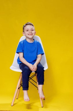 Cheerful little fair-haired boy in blue t-shirt and pants sitting on a chair and posing on yellow background.